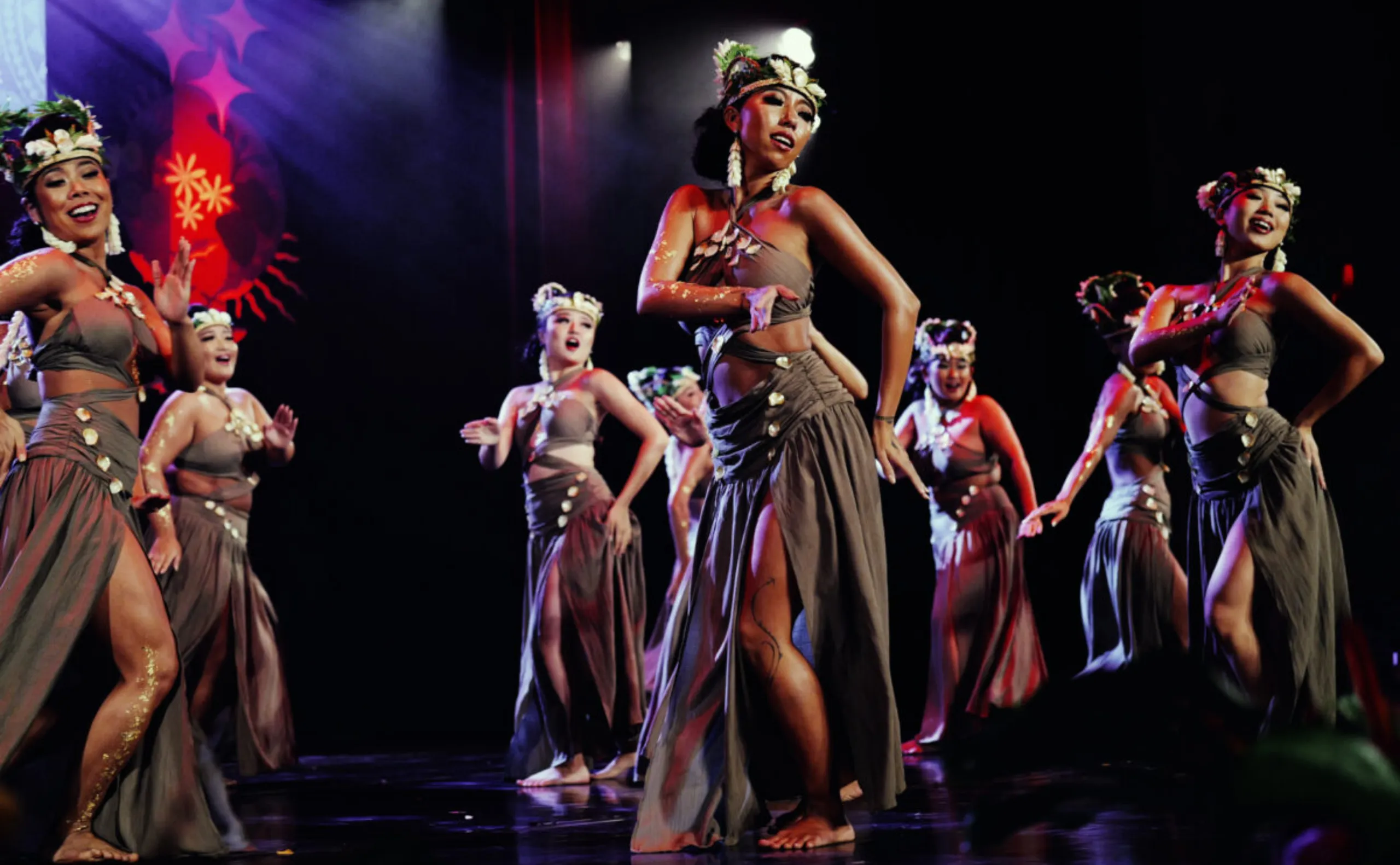 A group of women in traditional Polynesian attire perform a lively dance on stage, wearing brown skirts, floral headpieces, and jewelry, with expressive movements and joyful expressions under colorful stage lighting.