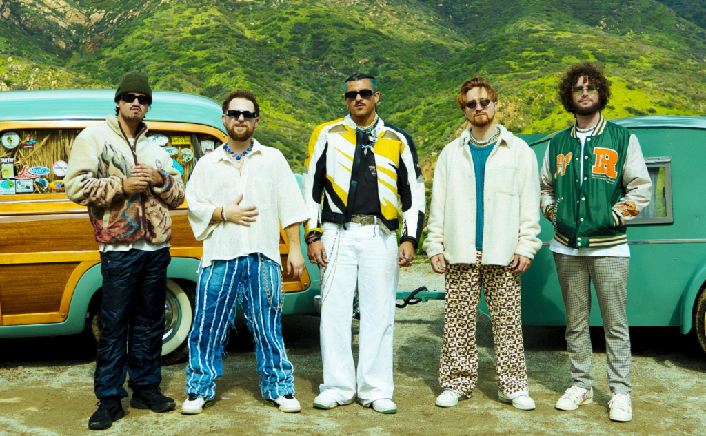 Five men stand in front of a vintage green car and trailer, wearing colorful, eclectic clothing, with green hills and mountains in the background.