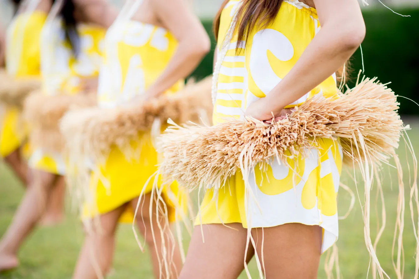 Women dressed in yellow and white costumes with raffia skirts stand outdoors in a line with hands on hips, preparing for or performing a traditional dance.