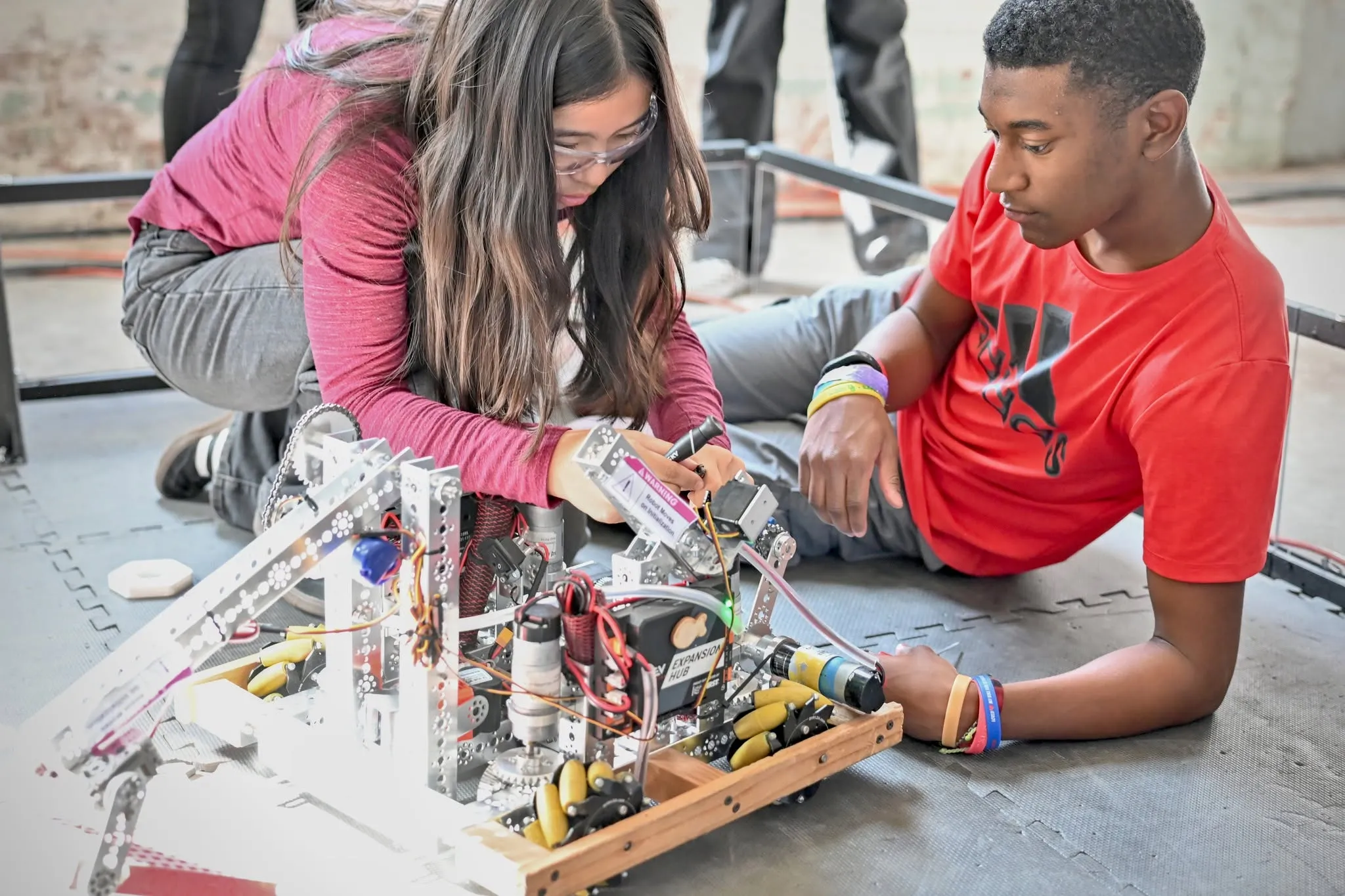 Two students work together on assembling or troubleshooting a complex robot, surrounded by mechanical parts and wires, while sitting on the floor inside a framed work area.