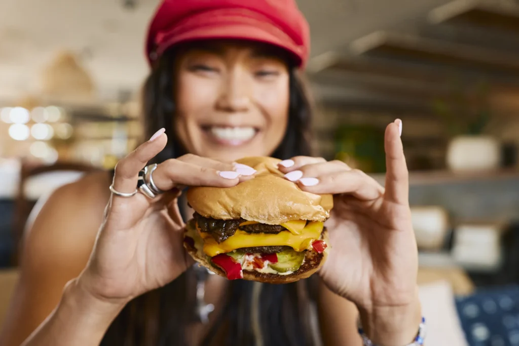 A person with long hair and a red hat smiles while holding a cheeseburger with toppings, shown up close in a casual indoor setting.