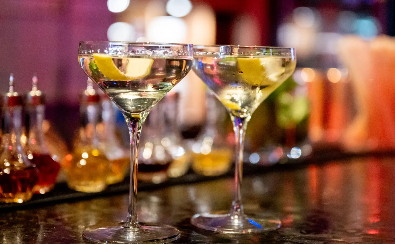 Two martini glasses with lemon twists on a dimly lit bar counter.