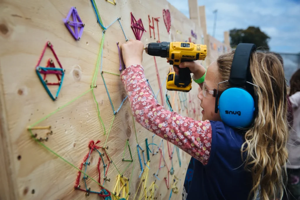 A young girl wearing safety earmuffs uses a cordless drill to attach colored string art to a wooden board. She is focused on her project, surrounded by other vibrant string designs.