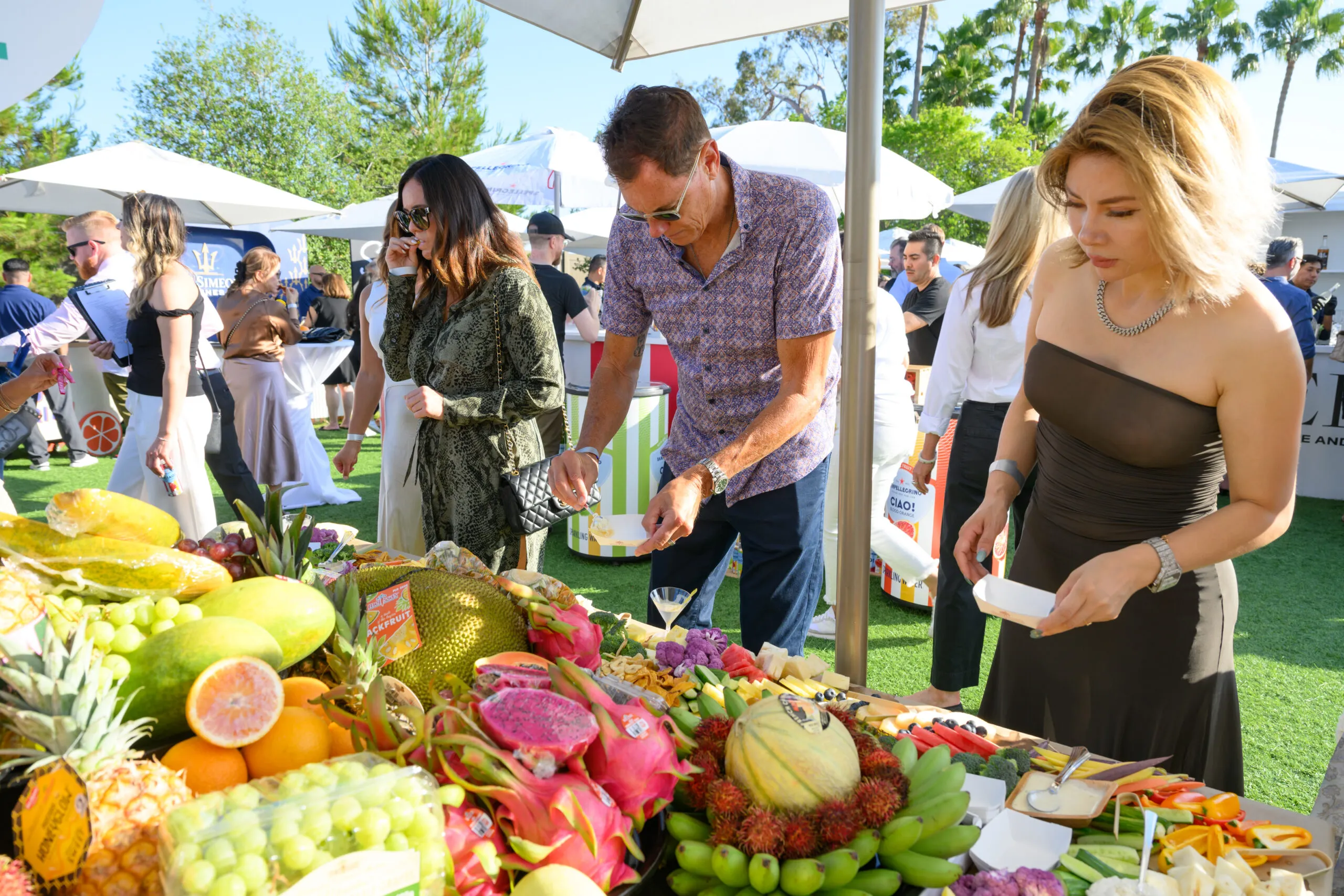 People gather at an outdoor event, serving themselves from a colorful tropical fruit buffet. Sunny weather, bright umbrellas, and palm trees create a lively, summery atmosphere.