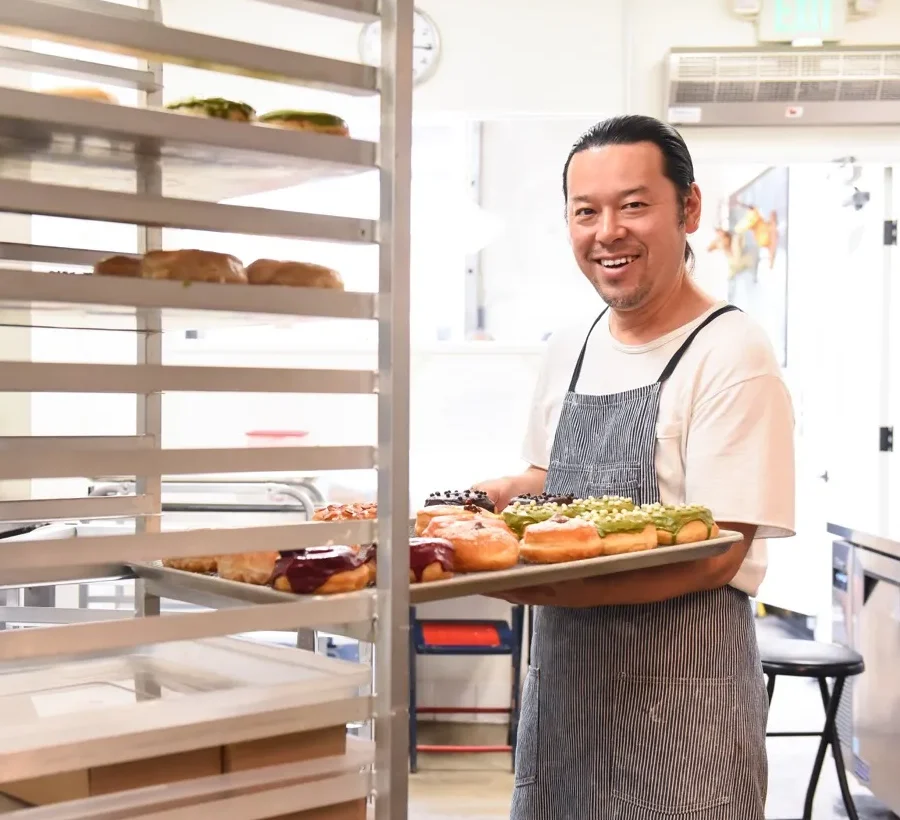 A smiling baker in an apron holds a tray of colorful donuts in a bright bakery kitchen, standing beside a metal cooling rack, celebrating the sweet traditions rooted in the history of Costa Mesa.