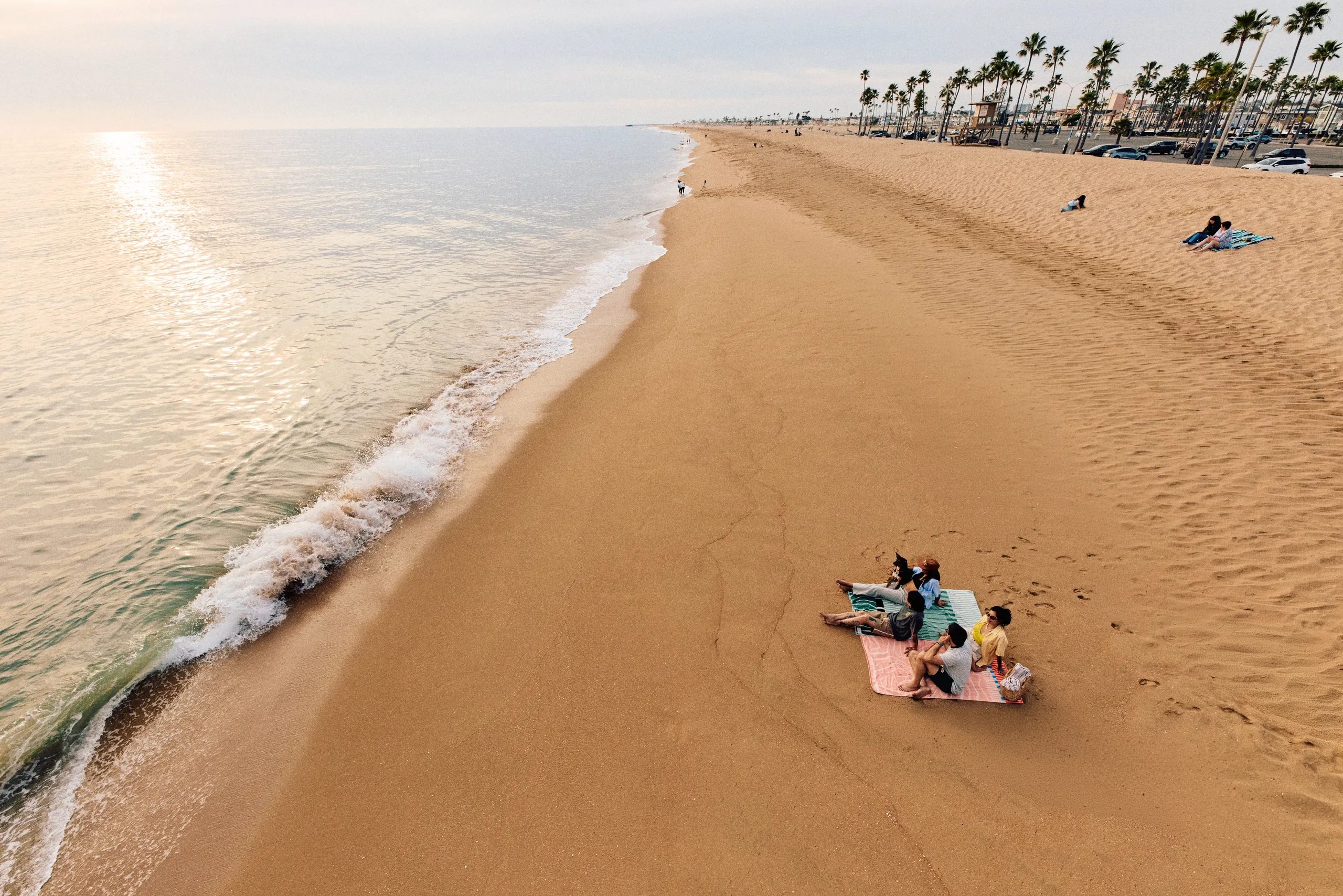 Three people relax on blankets on a wide sandy beach near the water, with gentle waves and palm trees lining the shore. The sun is low, creating a calm and peaceful atmosphere.