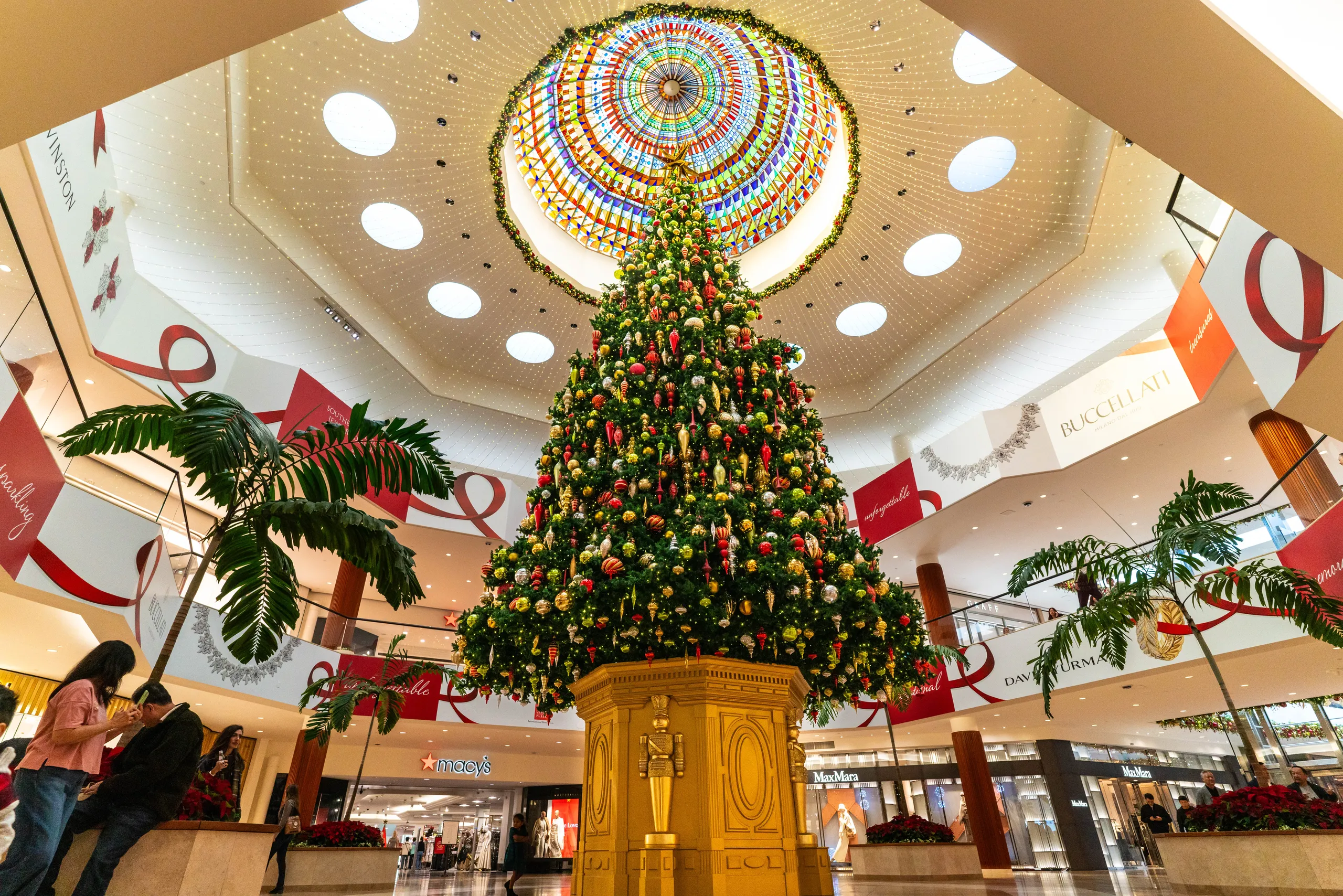 A large, decorated Christmas tree stands in the center of a shopping mall under a colorful, circular skylight, surrounded by shops and shoppers.