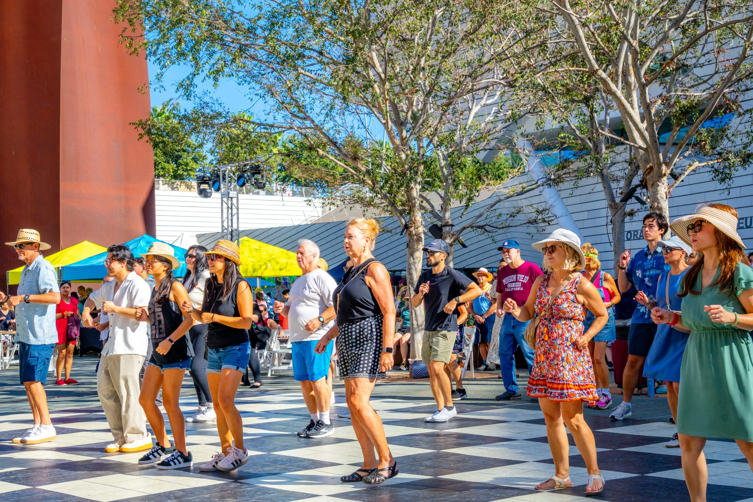 A group of people dances on a large outdoor checkered floor under trees, enjoying a sunny day in Costa Mesa. Some wear hats and sunglasses, as bright yellow tents highlight the lively history of Costa Mesa in the background.