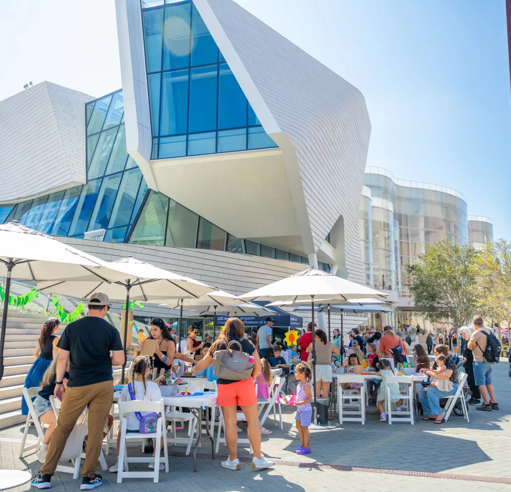People gather around tables with art supplies under umbrellas outside a modern, angular building on a sunny day. Children and adults participate in activities, creating a lively and friendly atmosphere that reflects the vibrant community spirit shaped by the history of Costa Mesa.