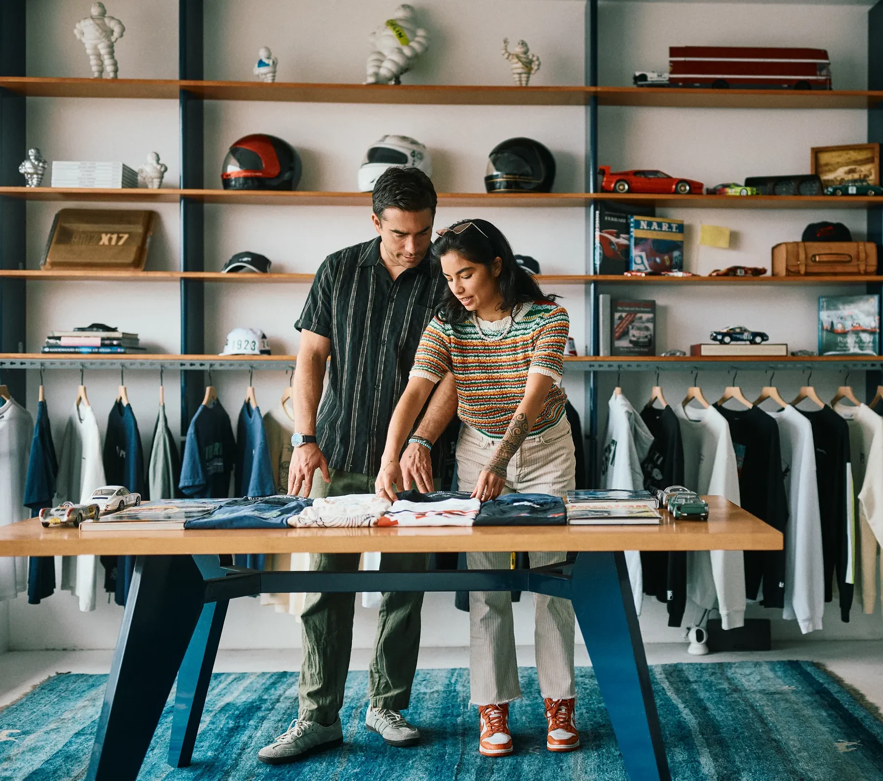 Two people stand at a table in a clothing store, examining shirts. Shelves behind them display hats, books, and model cars—some featuring nods to the unique history of Costa Mesa—while clothes hang neatly on a rack. The room has a modern, casual vibe with a blue rug.