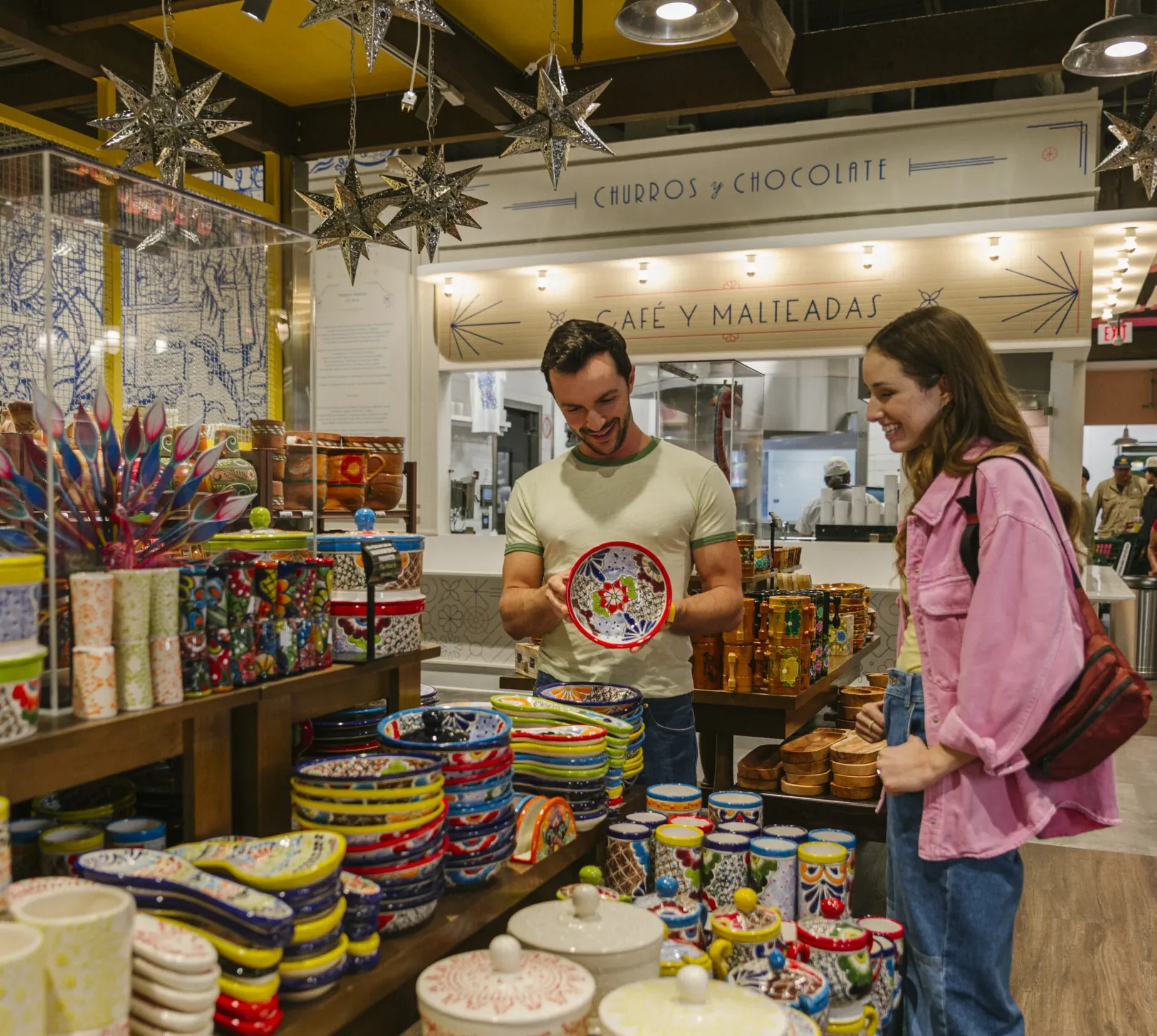 A man and woman browse colorful ceramic dishes at a vibrant market stall indoors, surrounded by decorative bowls, plates, and hanging star ornaments.