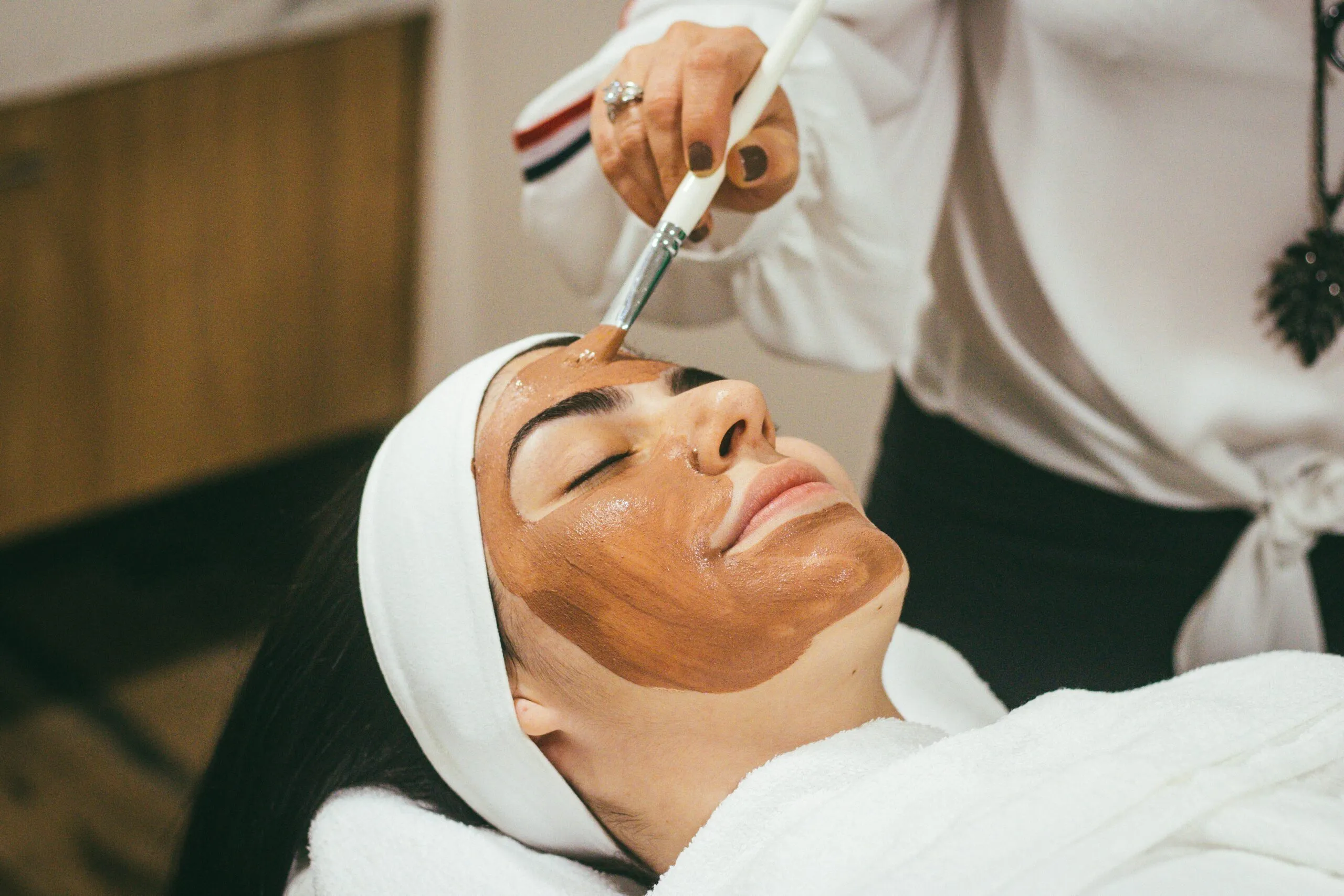 A person lying down with a white headband and towel, receiving a brown facial mask applied with a brush by another person.