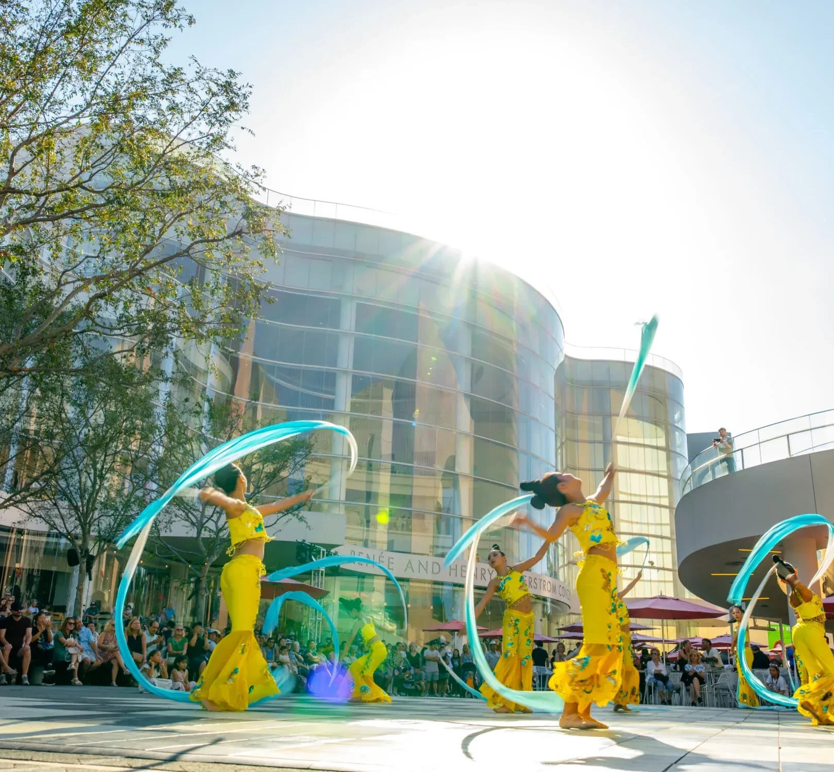 Four dancers in yellow costumes perform with flowing blue ribbons outdoors. A crowd watches, and the bright sun shines behind modern glass buildings and trees.