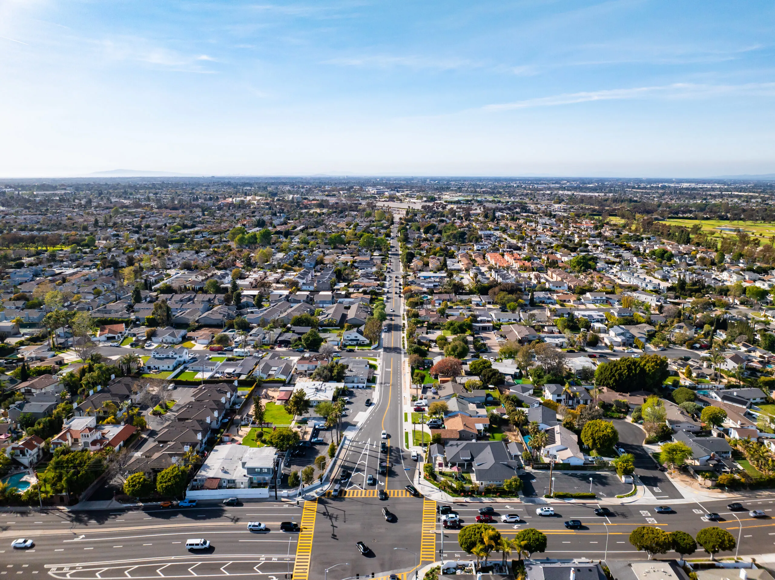 Aerial view of a suburban neighborhood in the history of Costa Mesa with grid-like streets, houses, trees, and cars at an intersection under a clear blue sky.