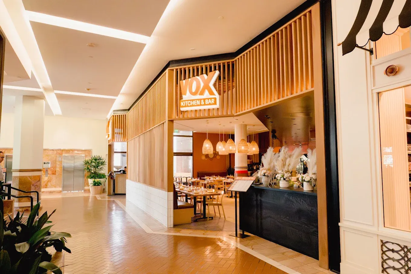 Vox Kitchen & Bar restaurant entrance in a mall, featuring wooden accents, hanging lights, indoor plants, and neatly arranged tables and chairs visible inside—offering a warm ambiance reminiscent of the inviting atmosphere found at Phoholic Costa Mesa.