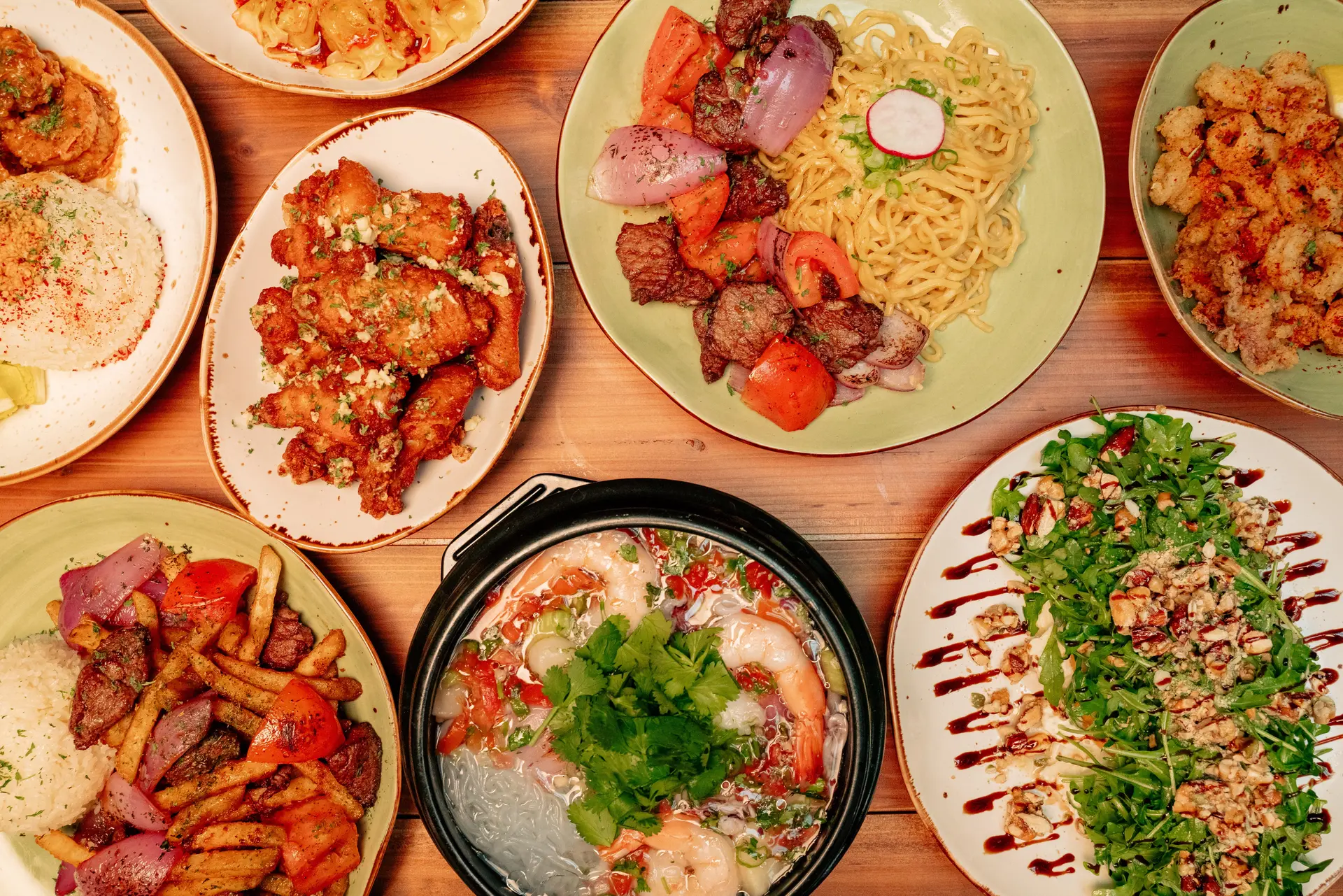 An overhead view of various dishes, including noodles, grilled meats, fried appetizers, salad, and a pot of soup with shrimp and vegetables, all arranged on a wooden table—reminiscent of the artful presentations found at Michelin star restaurants in Orange County.