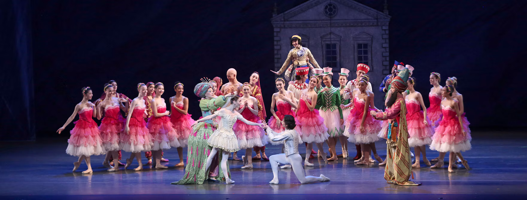 A large cast of ballet dancers in colorful holiday travel costumes performing on stage, with a male dancer kneeling to offer his hand to a ballerina in white.