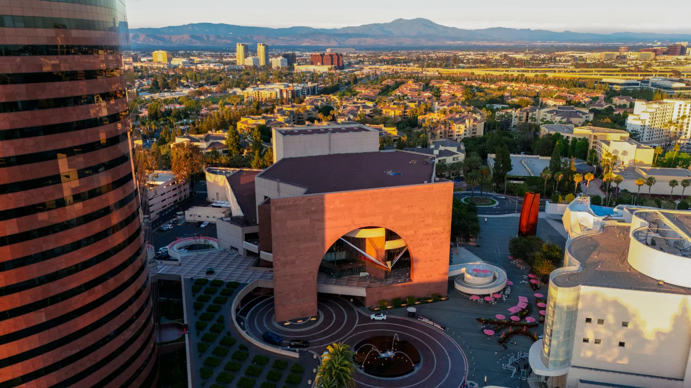 Aerial view of the Segerstrom Center for the Arts in Costa Mesa, featuring its arched building, plaza, and fountain, with urban sprawl and mountains in the distance, representing one of the many affordable things to do in the area.