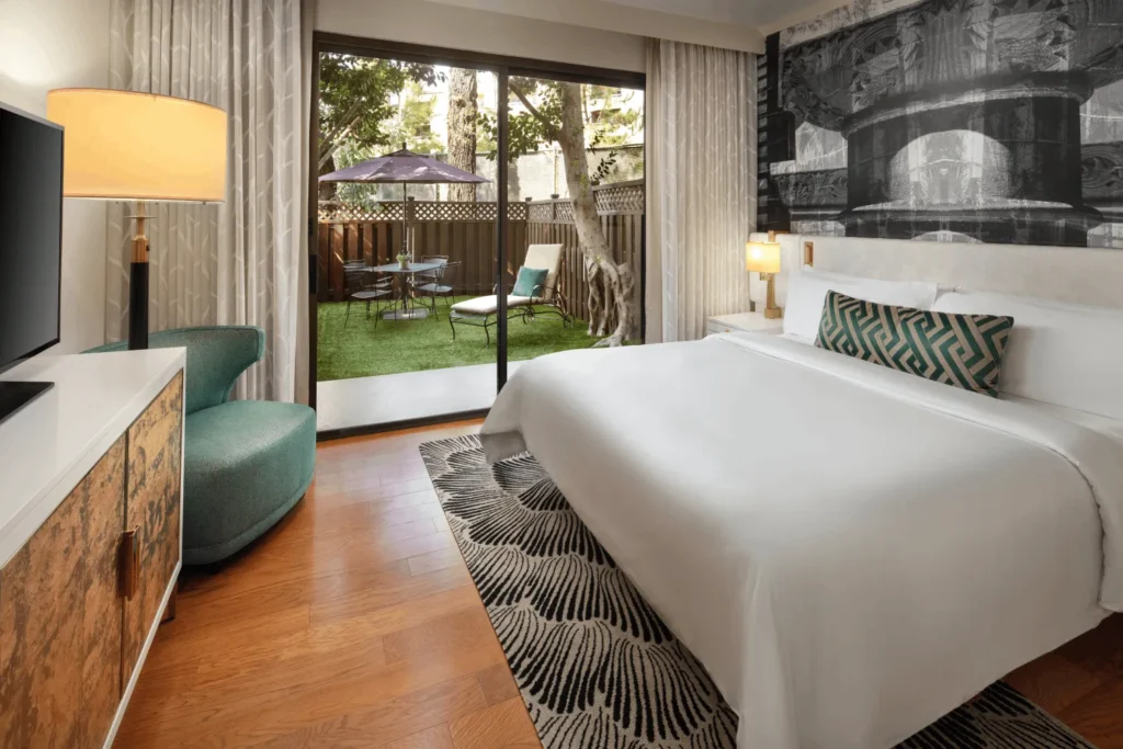 A modern hotel room with a large bed, patterned rug, green chair, TV, and sliding glass doors opening to a private patio with grass, trees, and outdoor seating.