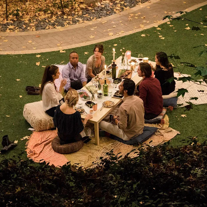 Six people seated on cushions around a low outdoor dining table on a lawn during an evening gathering, enjoying affordable things to do.