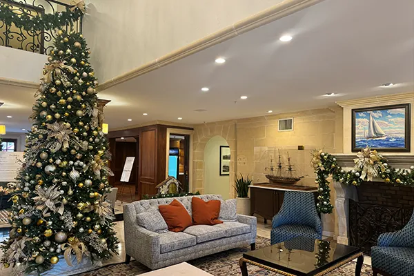 A holiday travel scene in a hotel lobby featuring a decorated Christmas tree near a fireplace, a grey sofa with orange throw pillows, and chairs adorned with garland.
