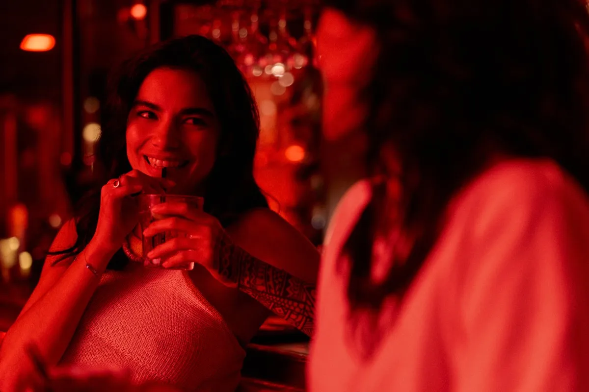 A woman with a tattooed arm smiles while drinking from a glass in a dimly lit bar with red lighting.
