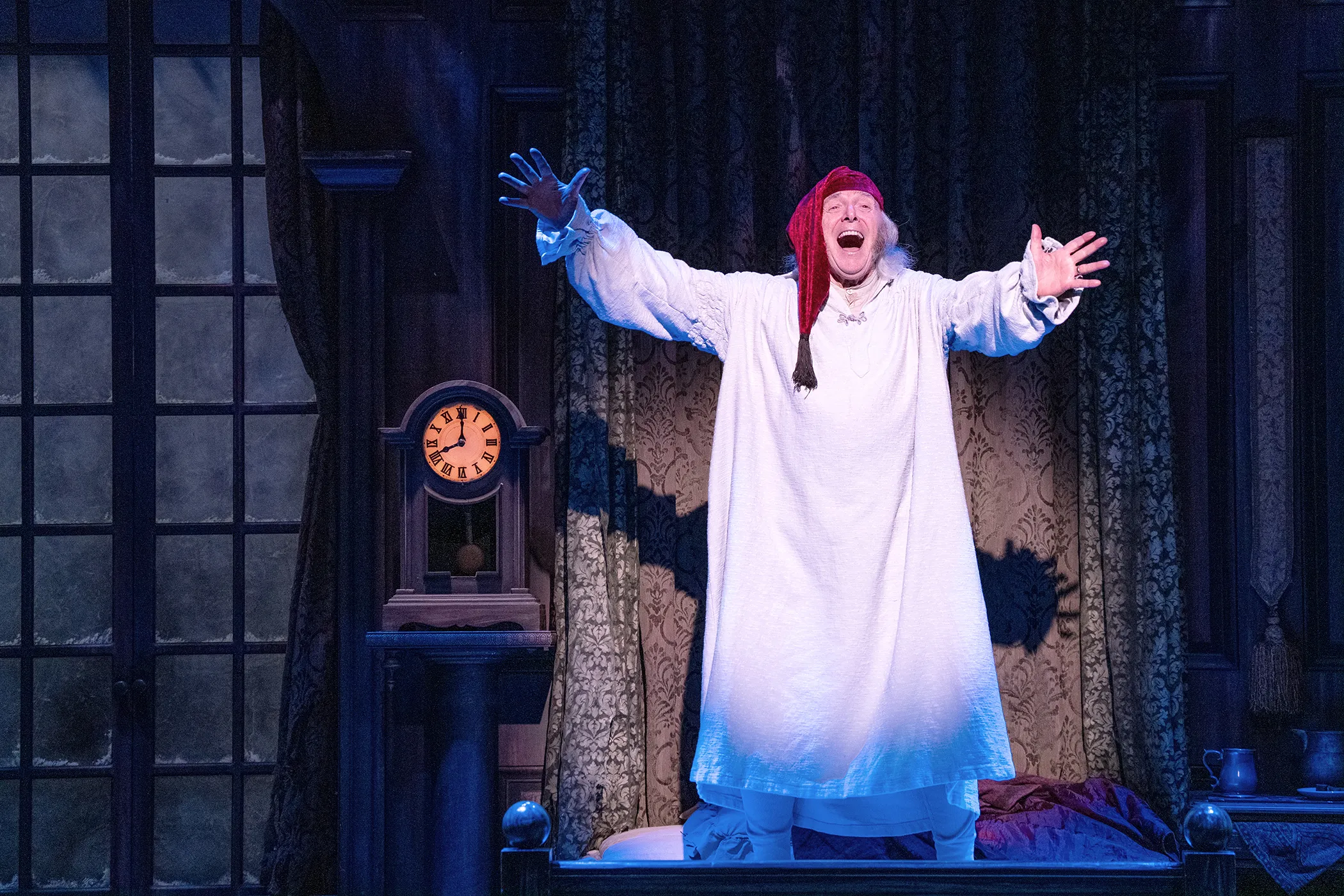 An actor playing Ebenezer Scrooge stands on a bed in a nightshirt and red nightcap, arms outstretched and mouth open in joy, embodying the spirit of holiday travel.