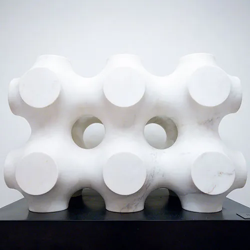 A white, modular marble sculpture on the Costa Mesa art walk featuring a geometric grid of interconnected spheres and hollow cylindrical openings, resting on a black pedestal.