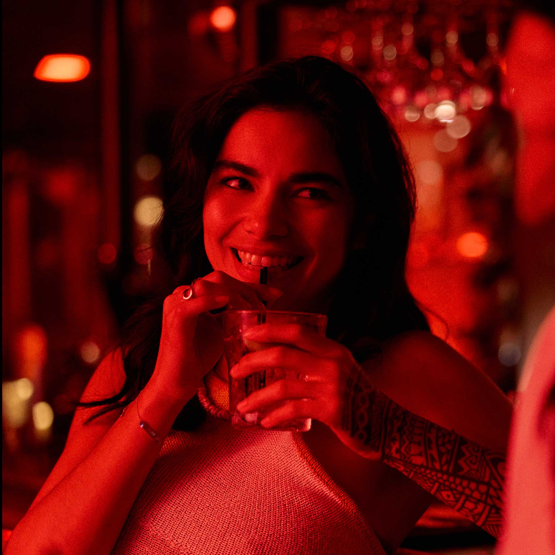 A woman with a patterned arm tattoo smiles while enjoying an affordable thing to do, drinking from a glass in a dimly lit, red-toned room.