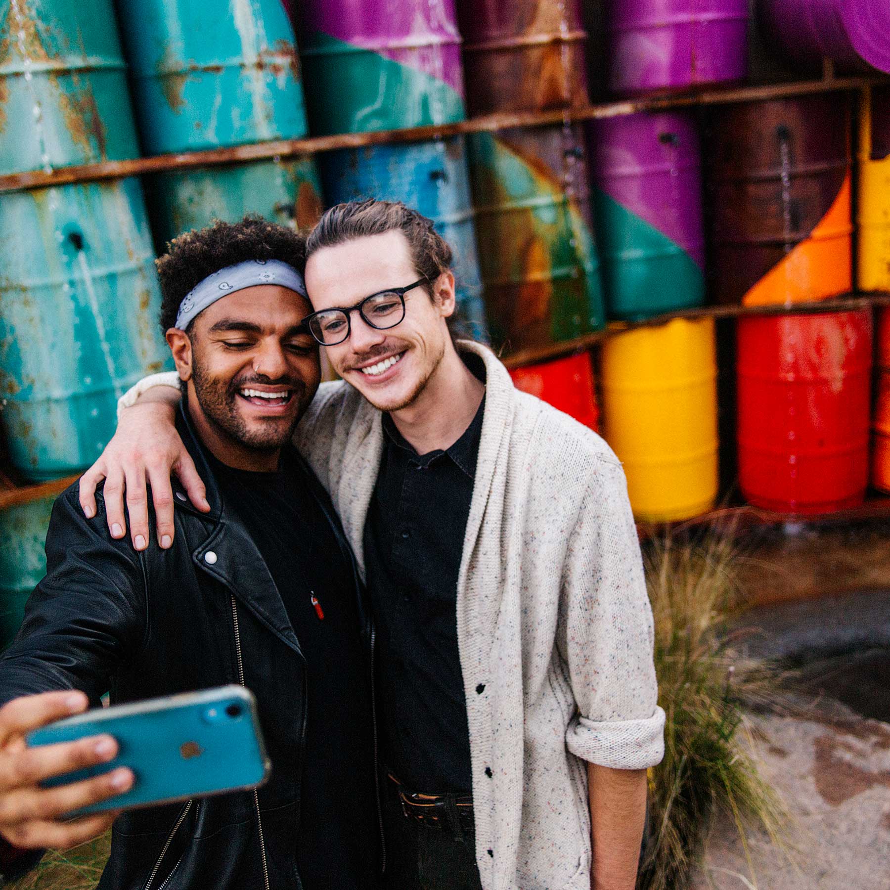 Two men smiling while taking a selfie together in front of a wall of colorful metal drums.