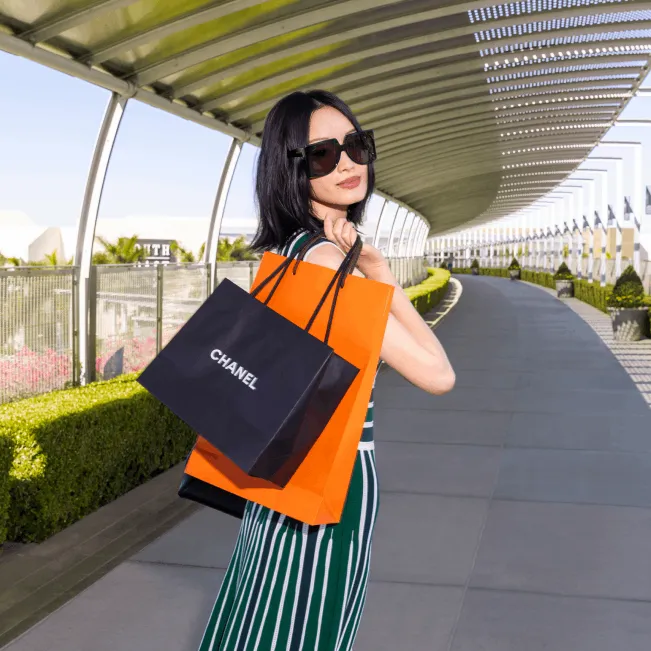 A woman in sunglasses and a green striped dress holds Chanel and orange shopping bags while walking outdoors on a sunny, modern walkway.
