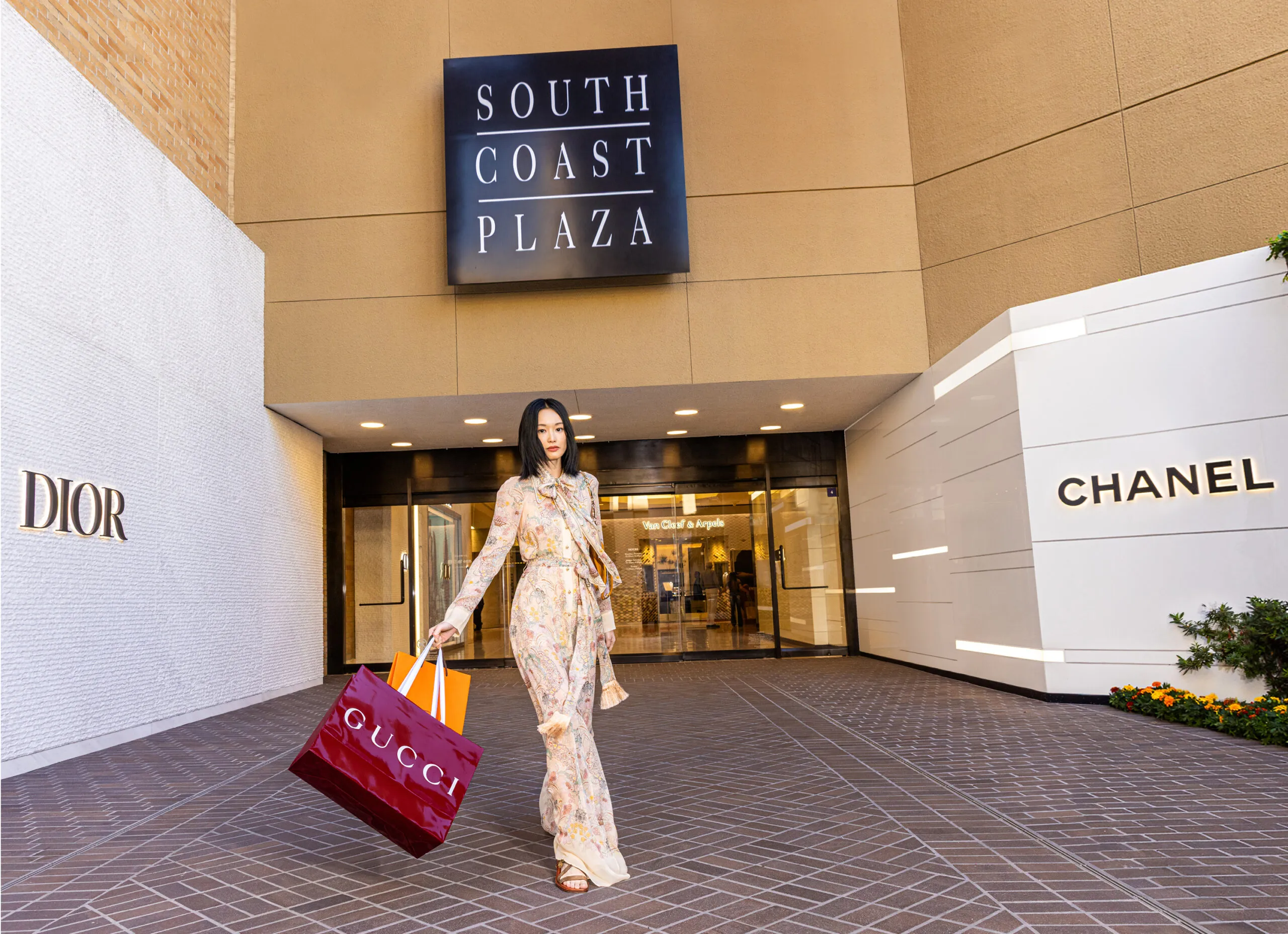 A woman in a floral dress carries Gucci shopping bags in front of the South Coast Plaza entrance, flanked by Dior and Chanel storefronts.
