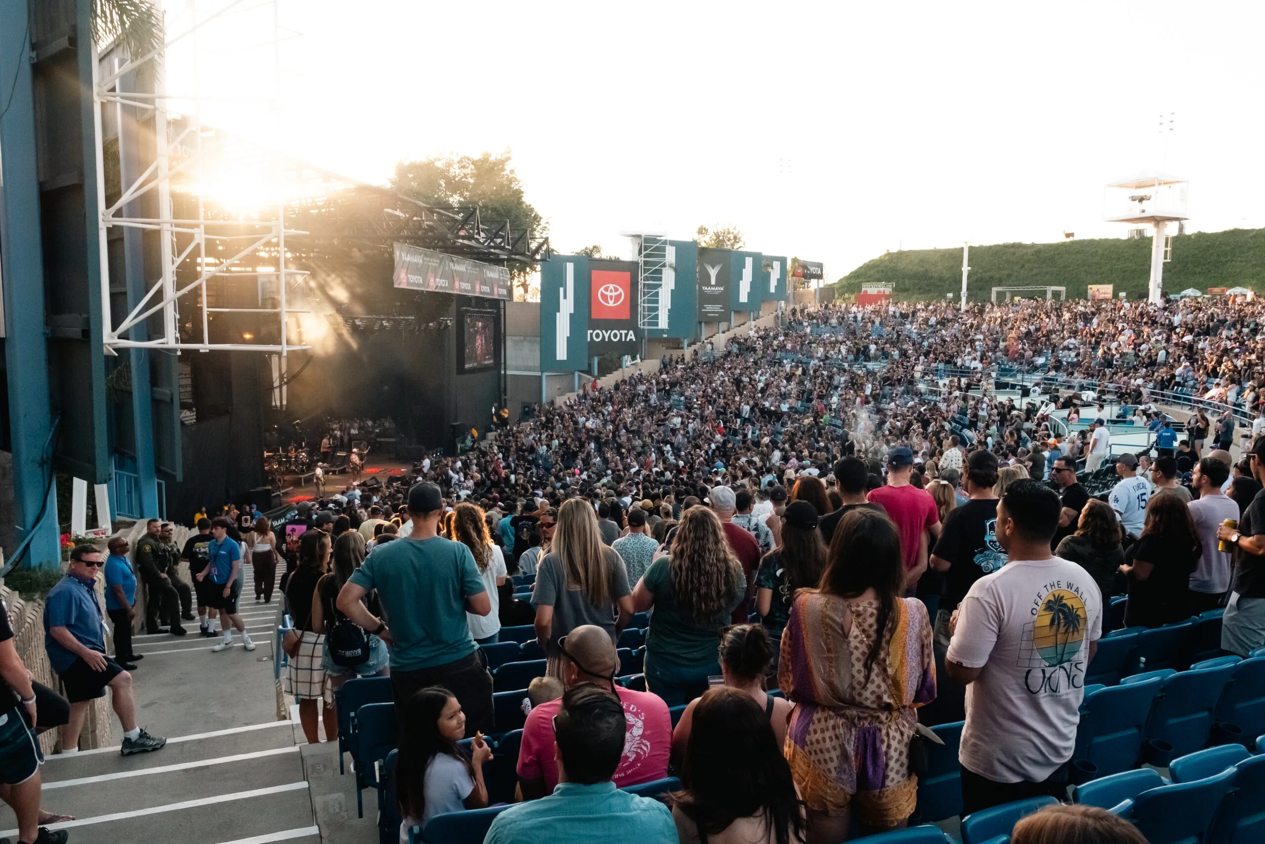 A large crowd fills an outdoor amphitheater, watching a concert as the sun sets—an event that boosts tourism economic impact as visitors enjoy the music near the stage, some standing and others in blue seats.