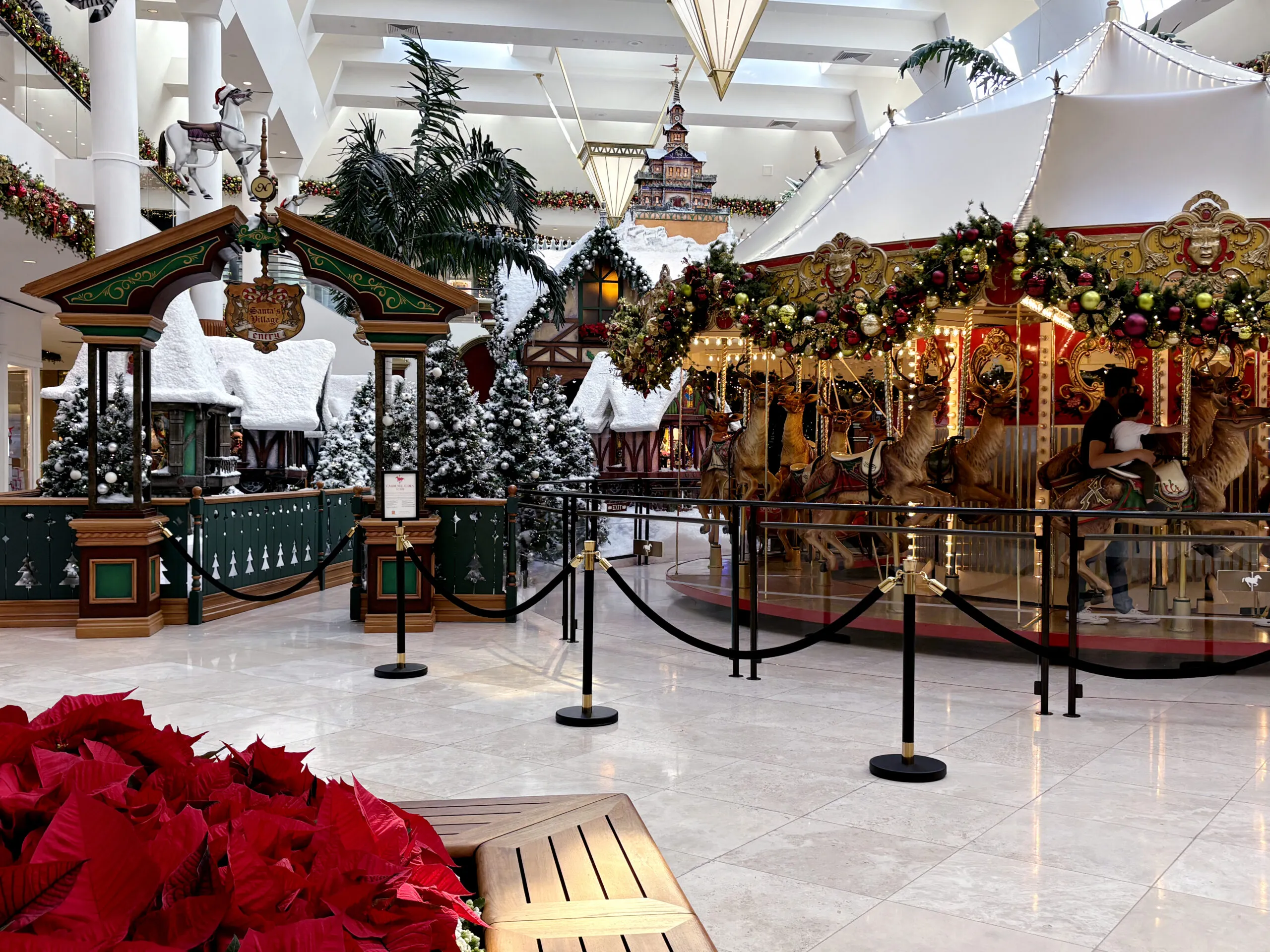 A holiday travel-inspired indoor display featuring a carousel with reindeer figures and a decorative archway, surrounded by snow-dusted evergreen trees and poinsettias.