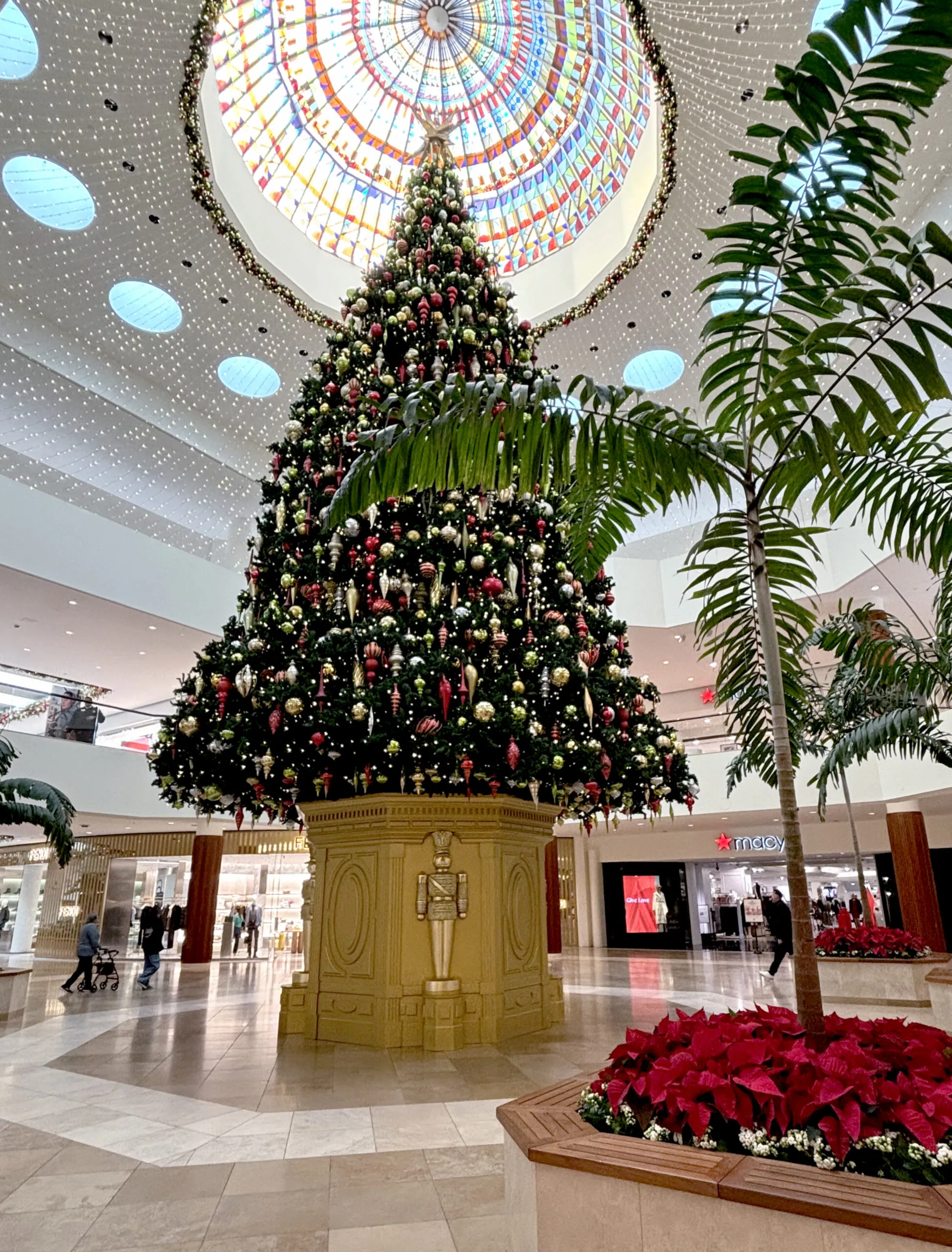 A large decorated Christmas tree stands in a shopping mall atrium beneath a colorful glass dome, creating a perfect spot for holiday travel, with a gold nutcracker base and potted red poinsettias in the foreground.