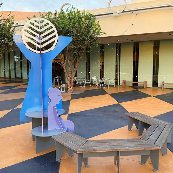 A blue metal sculpture featuring a circular top and a seated human figure, positioned on a patio along the Costa Mesa art walk near a curved wooden bench and outdoor seating.
