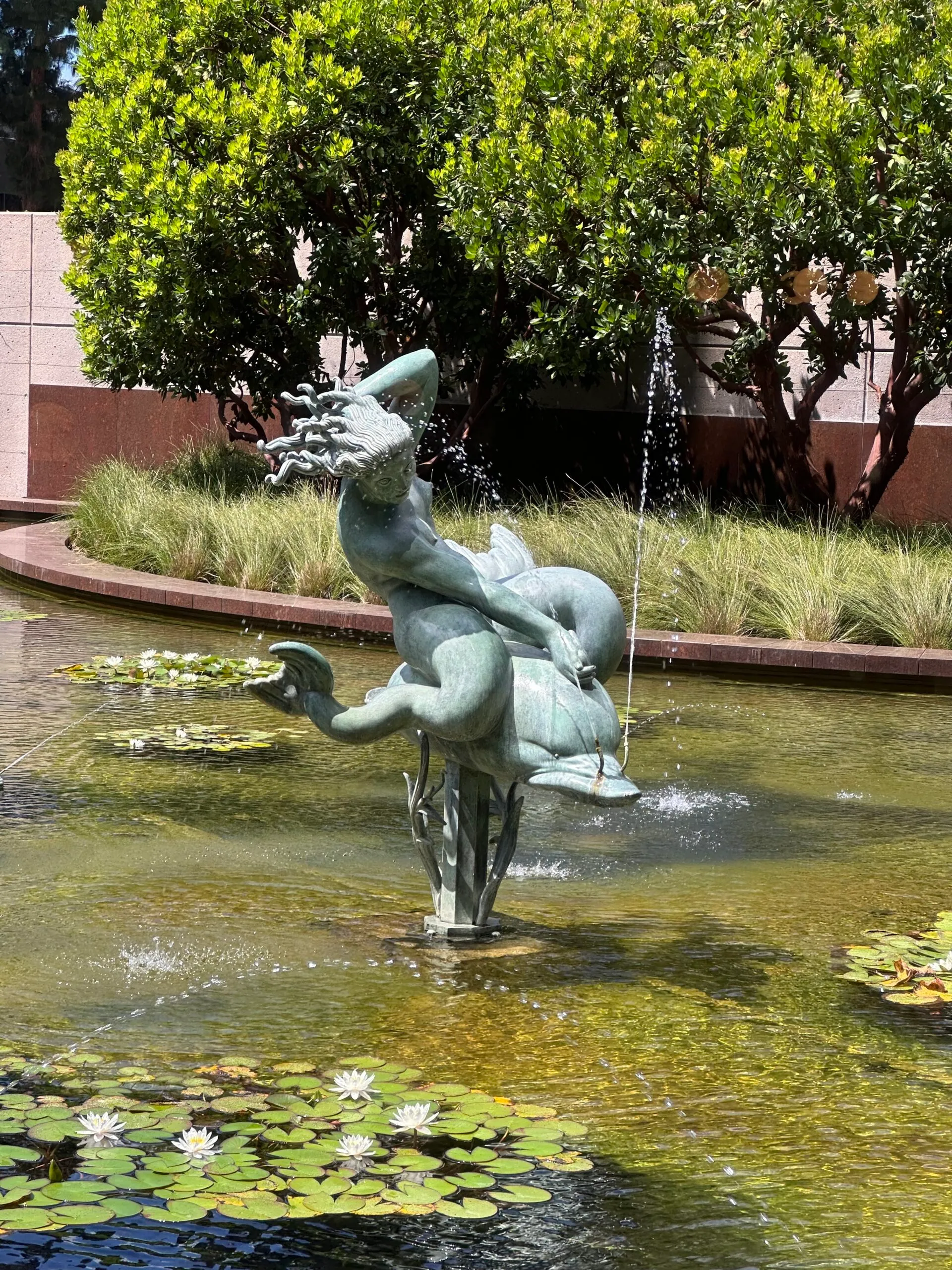 Bronze fountain of a figure riding a dolphin in the center of a pond filled with lily pads, a highlight of the Costa Mesa Art Walk.