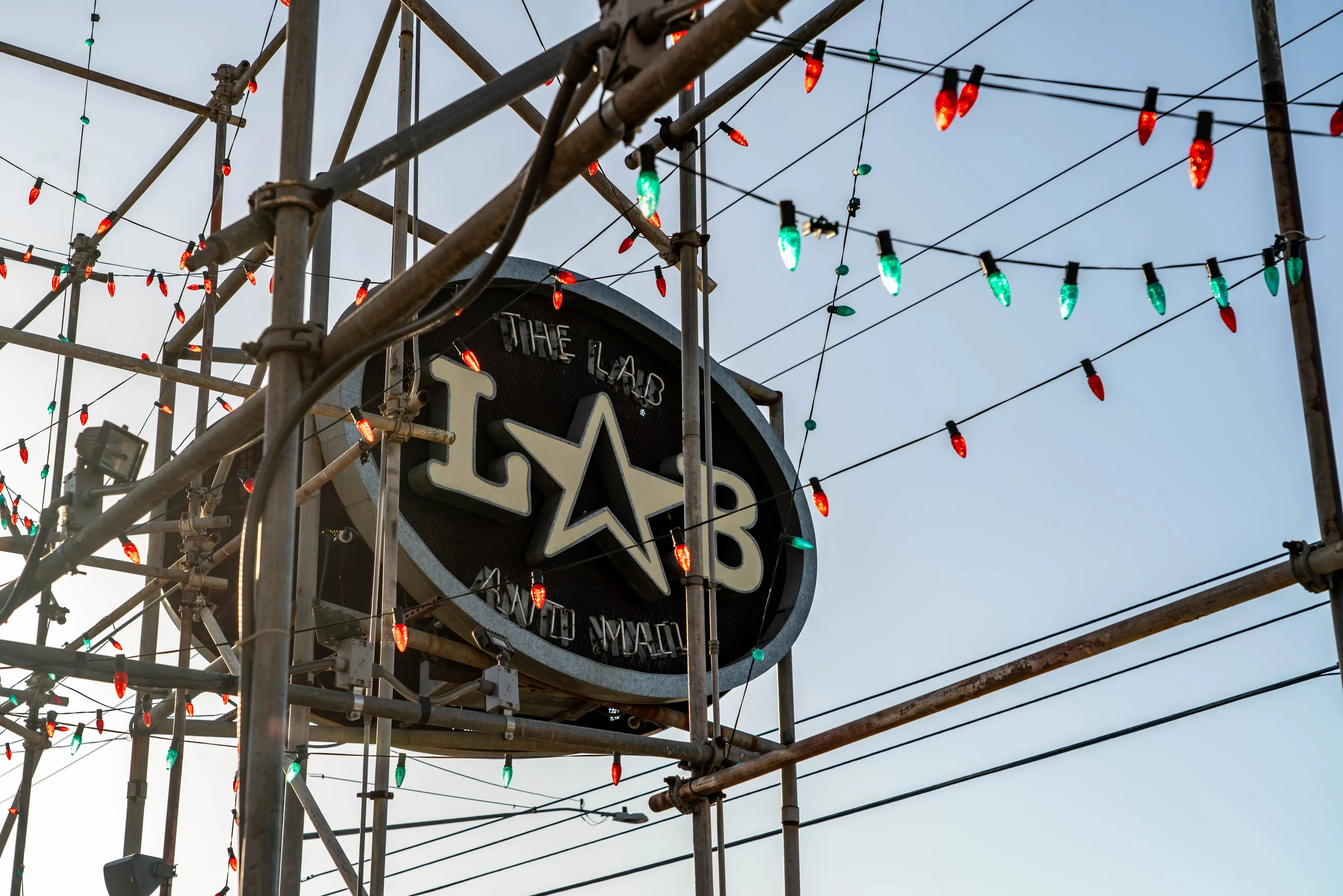 An oval THE LAB ANTI-MALL sign mounted on scaffolding, decorated with festive holiday travel lights against a blue sky.