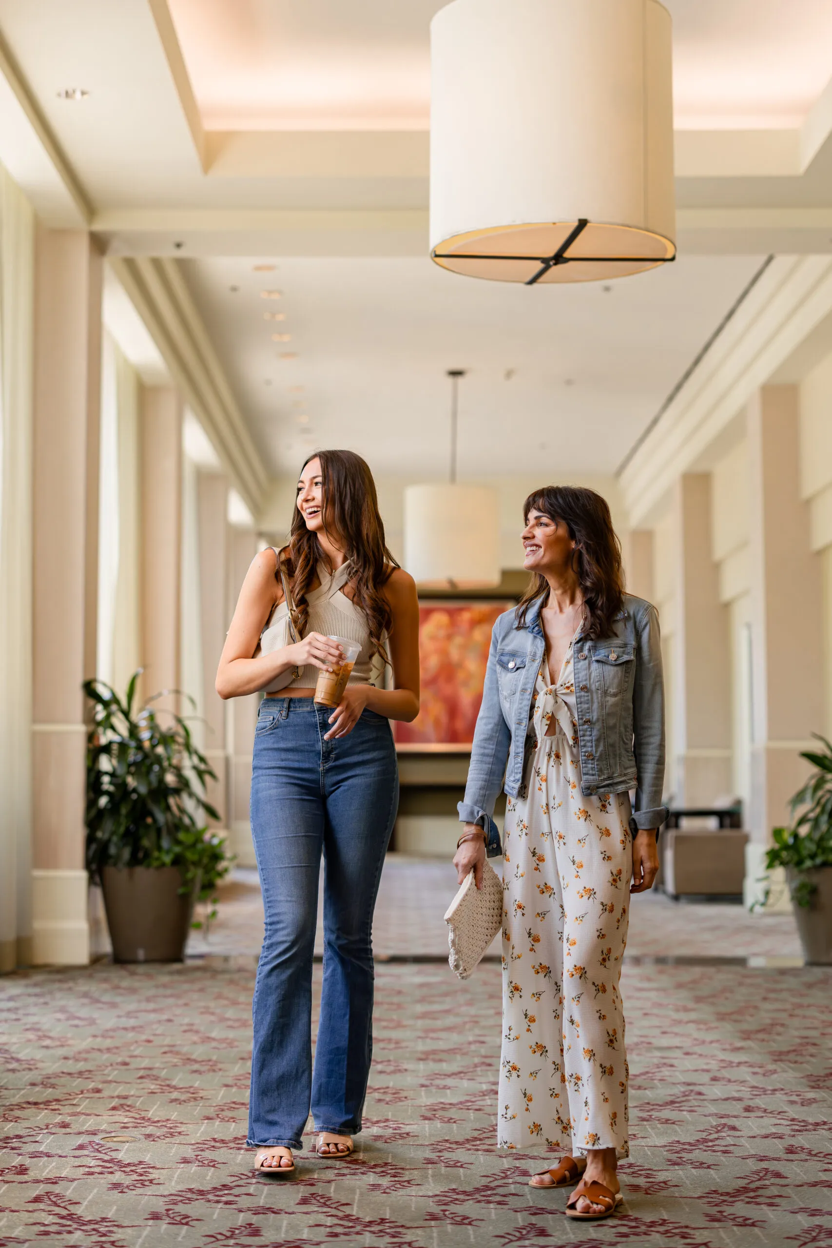 Two women walk and chat in a bright hallway. One wears jeans and a sleeveless top, holding coffee; the other wears a floral dress with a denim jacket, carrying a clutch. Large windows and plants line the modern space.