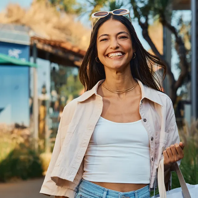 A smiling woman wearing a white tank top and unbuttoned shirt walks outdoors at a Costa Mesa shopping mall carrying shopping bags with sunglasses perched on her head.